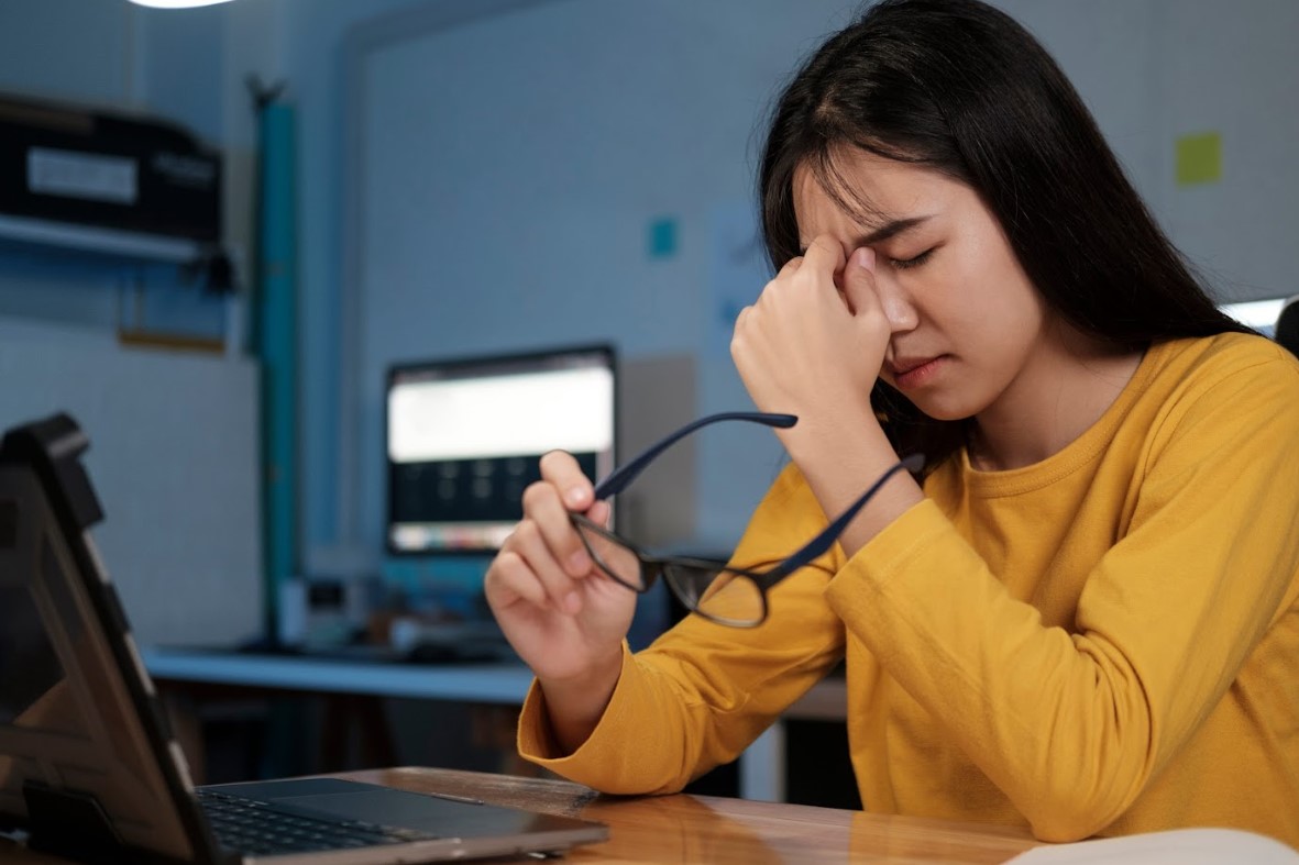Woman experiencing digital eye strain while working on a laptop, rubbing her eyes and holding glasses in a home office setting.